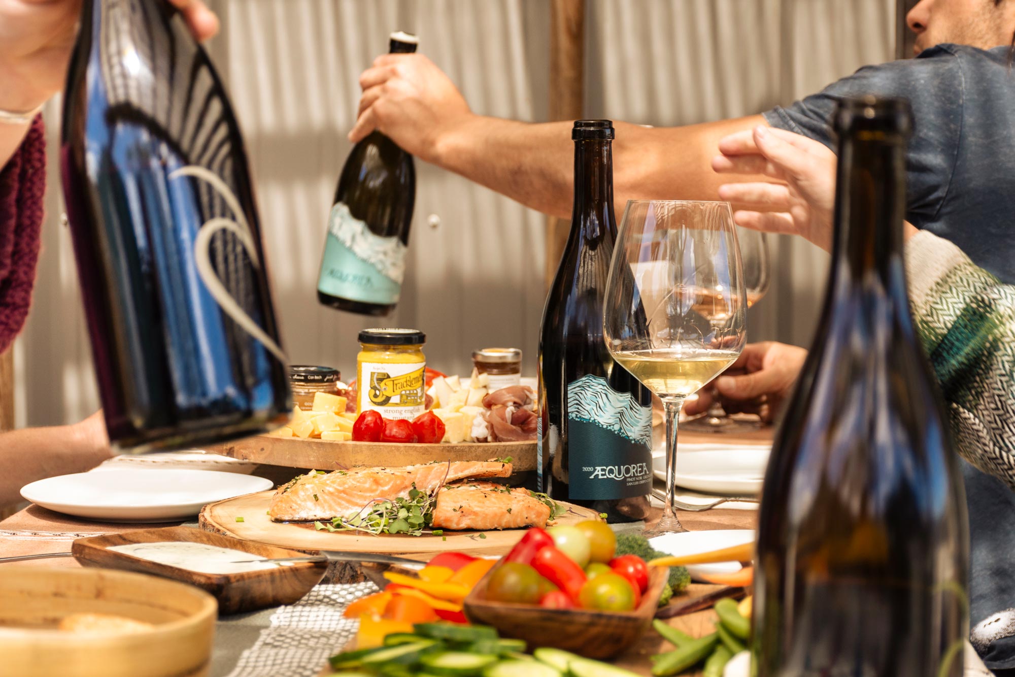 People passing wine bottles at a table full of appetizers and wine